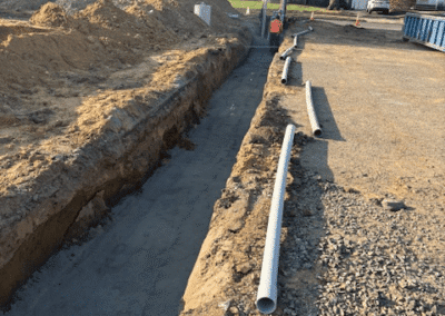 A construction site with a long trench dug in the ground, piles of dirt on the left, PVC pipes along the trench, and houses and trees visible in the background.