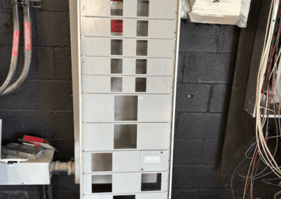 A tall, empty electrical panel with labeled slots is mounted on a black wall, surrounded by conduit pipes and exposed wires.
