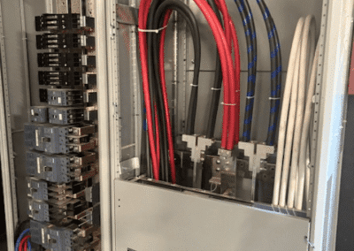 Electrical panel with thick red, black, and white power cables connected to bus bars, next to a compartment with multiple circuit breakers.
