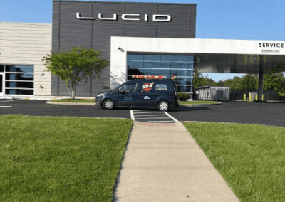 A sidewalk leads to a modern building with a large "Lucid" sign. A black van is parked in front of the entrance. The sky is clear and the grass is green.