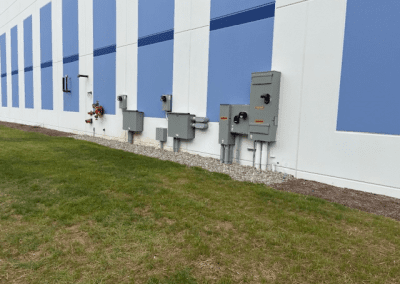 Electrical panels and utility boxes mounted on the exterior wall of a building with blue and white paneling, next to a grassy area.