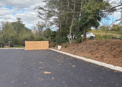 A newly paved parking lot with scattered pine needles, a wooden enclosure near the back, and trees lining the edge under a partly cloudy sky.