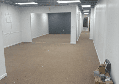 A hallway in an unfinished office space with exposed ceiling, white walls, brown carpet, and some boxes and items stacked in the corner.